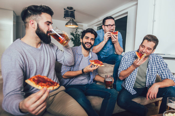Grupo de amigos varones positivos que beben cerveza y comen pizza en casa. - Foto, imagen