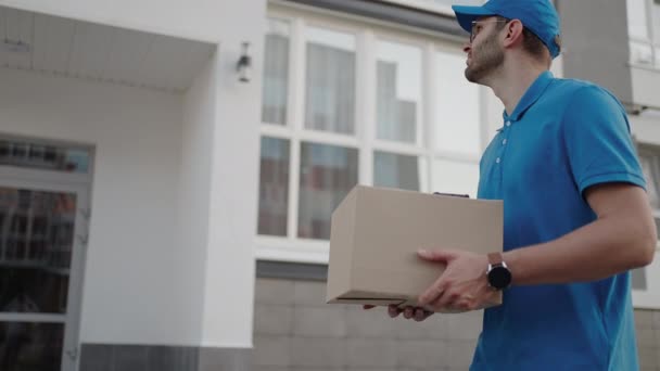 Молодой доставщик в очках и перчатках держит коробку с телефоном гуляет по карантину на улице торговый пакет пандемия самоизоляции почтальона замедленной съемки - Кадры, видео