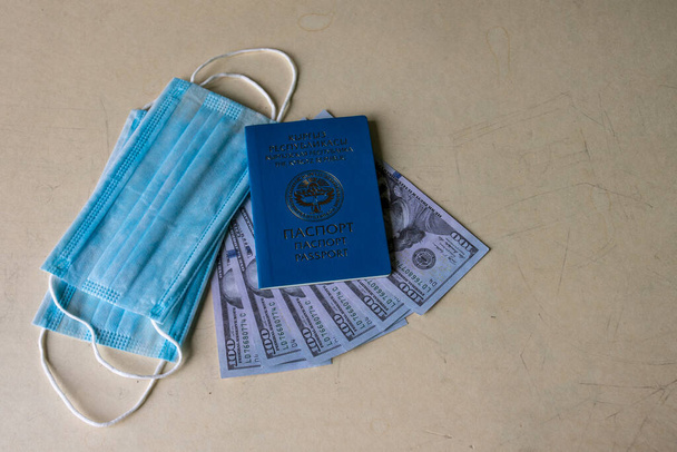 Ιατρική μάσκα, δολάρια, διαβατήριο Κιργιζίας. Διαβατήριο στα αγγλικά και ρωσικά, το οικόσημο της Κιργιζίας και η επιγραφή Δημοκρατία της Κιργιζίας. - Φωτογραφία, εικόνα