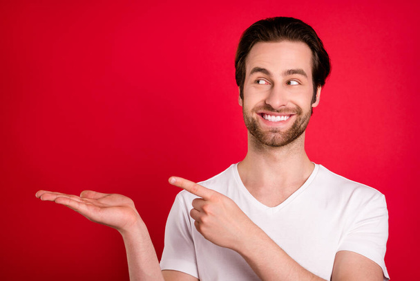 Photo of joyful young brown hairdo man hold point look empty space wear white t-shirt isolated on red color background - Fotoğraf, Görsel