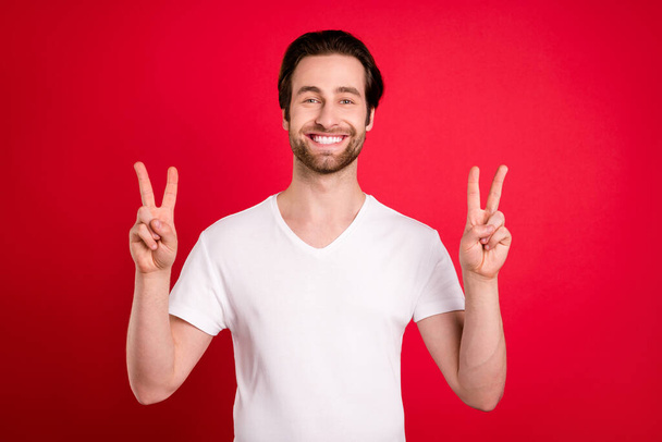 Photo of funny young brown hairdo man show v-sign wear white t-shirt isolated on vivid red color background - 写真・画像