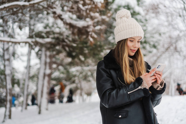 Happy nice girl using cellphone and smiling while walking in winter forest - 写真・画像