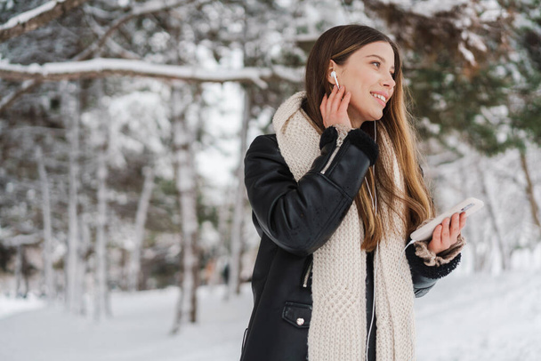 Happy girl in earphones using cellphone and smiling while walking in winter forest - Фото, изображение