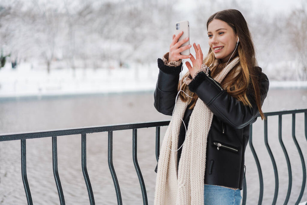 Szczęśliwa dziewczyna w słuchawkach robi zdjęcia na telefon komórkowy podczas spaceru w parku zimowym - Zdjęcie, obraz