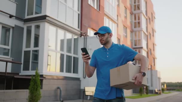 A male mail carrier with round glasses carries boxes for delivery to customers and looks at his mobile phone. Search through the mobile phone for the delivery address. Payment terminal. - Footage, Video