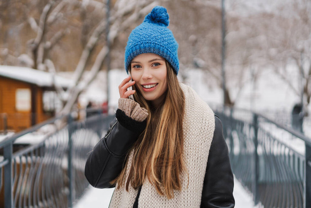 Beautiful happy young woman in winter jacket standing on a bridge, using mobile phone - Foto, Imagem