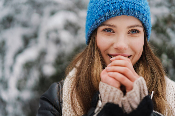 Happy beautiful girl in knit hat warming her hands while walking in winter forest - Fotografie, Obrázek