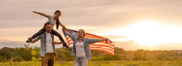 Fiatal szülők lányukkal, akik amerikai zászlót tartanak vidéken naplementekor. Függetlenség Napja ünneplés - Fotó, kép