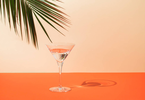 Top view of green tropical palm leaf and martini glass on orange and sand color background. Summer drink minimal concept. Elegant composition. - Photo, image
