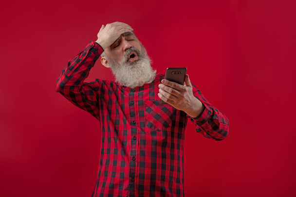 Ενηλίκων όμορφος ανώτερος αρσενικό μοντέλο με γκρι γενειάδα φορώντας καρό κόκκινο και μαύρο πουκάμισο δείχνει σοκαρισμένος facepalm έκπληξη, δυσαρέσκεια, κοιτάζοντας το smartphone - Φωτογραφία, εικόνα
