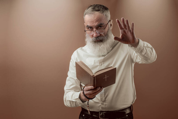Hombre carismático mayor con una barba gris en una camisa ligera sosteniendo el libro de la Santa Biblia y predicando - Foto, imagen