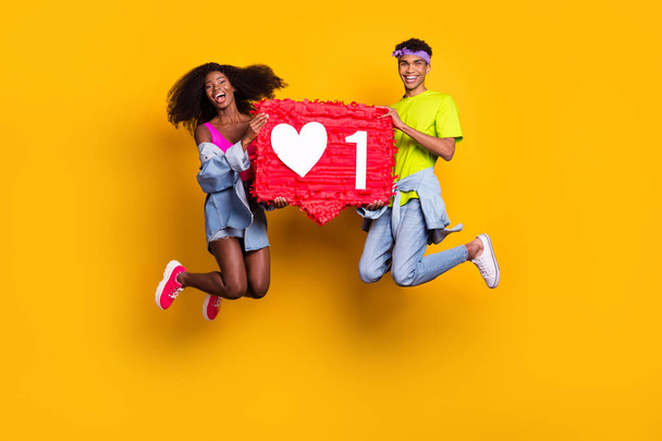 Full length body size view of handsome beautiful trendy couple jumping holding red board isolated on bright yellow color background - Fotó, kép