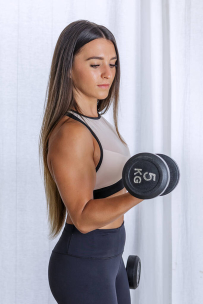 Vista laterale di una forte atleta donna che fa riccioli di bicipiti con manubri pesanti durante l'allenamento a casa - Foto, immagini