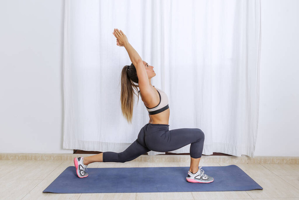 Visão lateral do corpo inteiro da fêmea apto em sportswear e tênis de pé em Crescent posição lunge com os braços levantados durante o alongamento do corpo durante o treinamento de ioga fitness em casa - Foto, Imagem