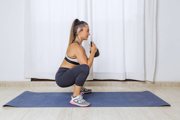 Boční pohled plné tělo fit mladé ženy ve sportovním oblečení squatting s kettlebell při tréninku svaly při fitness cvičení doma - Fotografie, Obrázek
