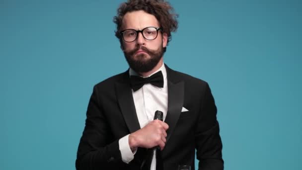 proud happy bearded guy in black tuxedo with eyeglasses holding champagne and glasses, nodding and recommending wine bottle, smiling and having fun on blue background in studio - Filmati, video
