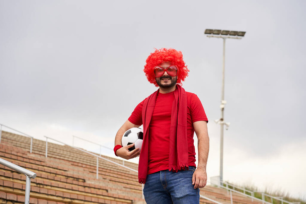 Fotbalový fanoušek s parukou a klaunskými brýlemi na stadionu se dívá do kamery a drží míč. - Fotografie, Obrázek