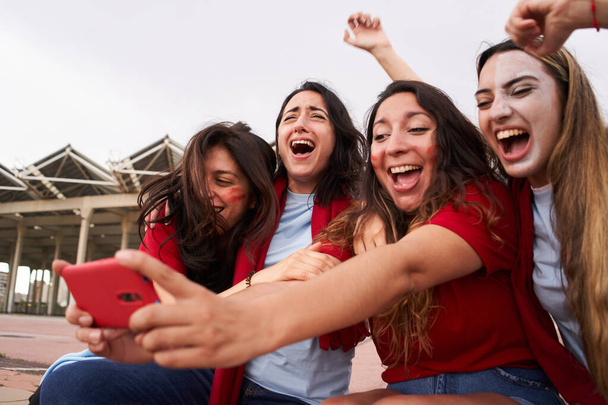 Grupo de seguidoras de un equipo de fútbol viendo un partido en streaming vestidas con camisetas rojas. Las mujeres celebran alegremente el gol con la euforia. - Foto, imagen