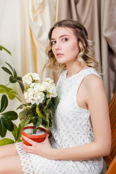 Retrato de uma bela jovem mulher em um vestido branco com um pote de flores brancas artificiais em suas mãos sentadas em uma cadeira contra um fundo de drapejamento bege claro. - Foto, Imagem