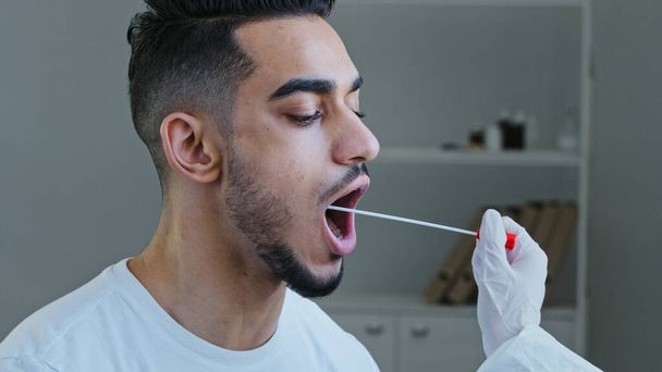 Primo piano uomo spagnolo arabo maschio paziente apre bocca dona campione di saliva per il virus dell'infezione, infermiere medico irriconoscibile facendo test PCR con attrezzature per covid-19 coronavirus al ragazzo etnico - Foto, immagini