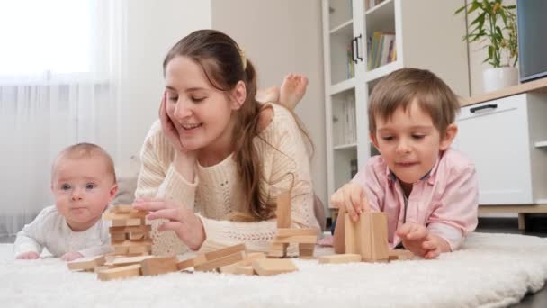Pequeño bebé, madre y hermano mayor jugando con bloques de madera en el suelo en la sala de estar. Crianza, felicidad de los hijos y relación familiar - Metraje, vídeo
