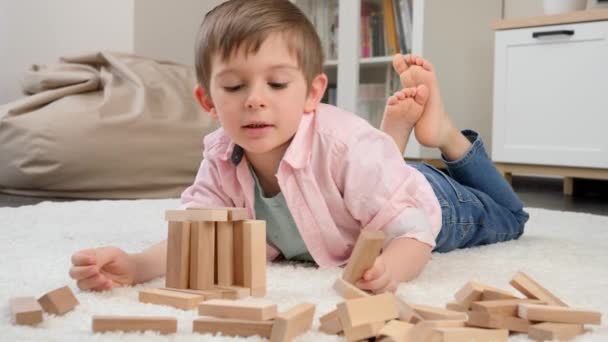 Милый маленький мальчик лежит на ковре и играет с игрушечными деревянными блоками. Концепция детского образования и игр на дому - Кадры, видео
