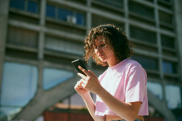 Милая молодая женщина сидит на улице в наушниках и выбирает хорошую музыку на телефоне. - Фото, изображение