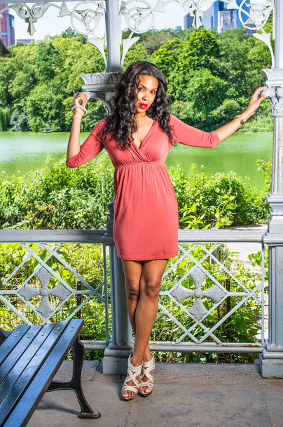 Dressing in a red dress, a pretty black woman with long curly hair is standing by a pavilion in a park, charmingly looking at you - Foto, afbeelding