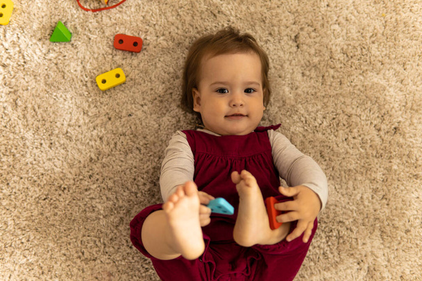 care, childhood - above view portrait one little happy funny smiling authentic child baby toddler kid in pink have fun hug legs play with shapes lay on soft white floor mat indoors at cozy sweet home - Фото, изображение