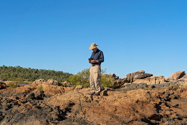 Кейп Глостер, Квінсленд, Австралія - червень 2021: чоловік - пенсіонер стоїть на скелях, утримуючи контроль над дроном під час польоту над ландшафтом. - Фото, зображення