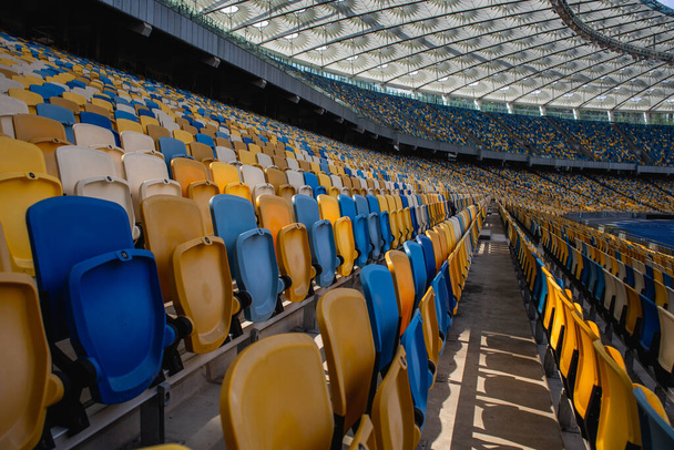 Prázdné řady sedadel na fotbalovém olympijském stadionu se žlutými a modrými lavicemi - Fotografie, Obrázek