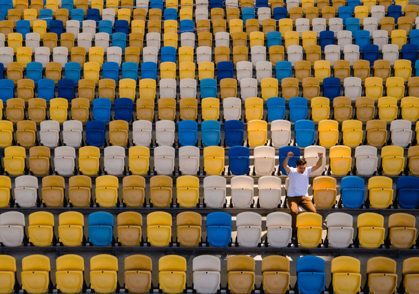 Un joven con una camiseta blanca está cantando en una tribuna vacía de un estadio de fútbol - Foto, imagen