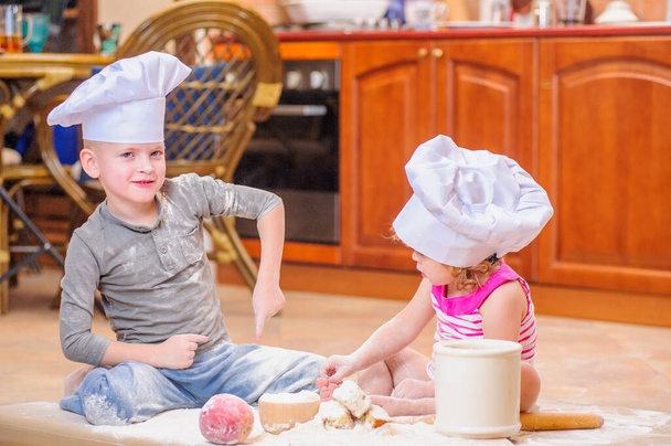 Dwoje rodzeństwa - chłopiec i dziewczynka - w czapkach szefa kuchni przy kominku, siedzącym na podłodze w kuchni zabrudzonym mąką, bawiącym się jedzeniem, robiącym bałagan i bawiącym się - Zdjęcie, obraz