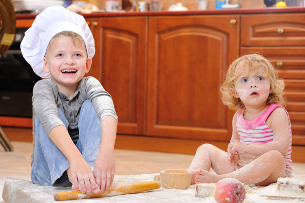 Deux frères et sœurs - garçon et fille - dans les chapeaux du chef près de la cheminée assis sur le sol de la cuisine souillé avec de la farine, jouer avec la nourriture, faire du désordre et s'amuser - Photo, image
