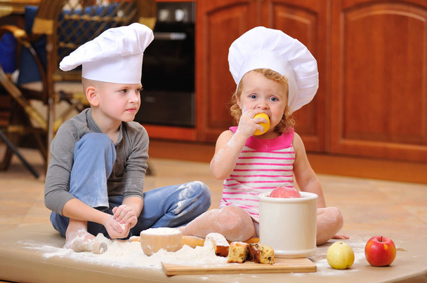 Zwei Geschwister - Junge und Mädchen - in Kochmützen neben dem Kamin sitzen auf dem mit Mehl beschmutzten Küchenboden, spielen mit Essen, machen Chaos und haben Spaß - Foto, Bild