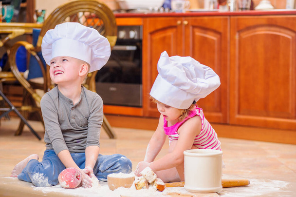 Dois irmãos - menino e menina - em chapéus de chef perto da lareira sentados no chão da cozinha sujos de farinha, brincando com comida, fazendo bagunça e se divertindo - Foto, Imagem