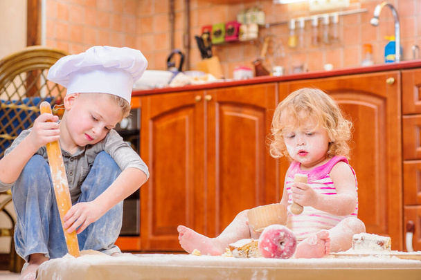 Kaksi sisarusta - poika ja tyttö - keittiömestarin hatuissa lähellä takkaa, istuen keittiön lattialla jauhoilla likaantuneena, leikkien ruoalla, tehden sotkua ja pitäen hauskaa - Valokuva, kuva