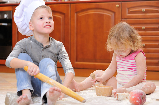 2人の兄弟-男の子と女の子-台所の床に座っている暖炉の近くのシェフの帽子に小麦粉で汚れ、食べ物と遊び、混乱を作り、楽しみを持っています - 写真・画像