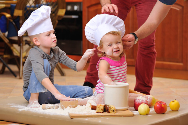 Dva sourozenci - chlapec a dívka - v kuchařských čepicích u krbu, sedící na podlaze v kuchyni poskvrněné moukou, hrající si s jídlem, dělají nepořádek a baví se - Fotografie, Obrázek