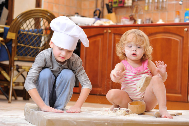 Dos hermanos - niño y niña - en sombreros de chef cerca de la chimenea sentados en el suelo de la cocina manchados de harina, jugando con la comida, haciendo lío y divirtiéndose - Foto, Imagen