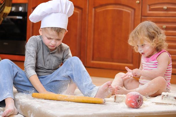 Δύο αδέλφια - αγόρι και κορίτσι - με καπέλα σεφ κοντά στο τζάκι κάθονται στο πάτωμα της κουζίνας λερωμένα με αλεύρι, παίζοντας με το φαγητό, κάνοντας χάος και διασκεδάζοντας - Φωτογραφία, εικόνα