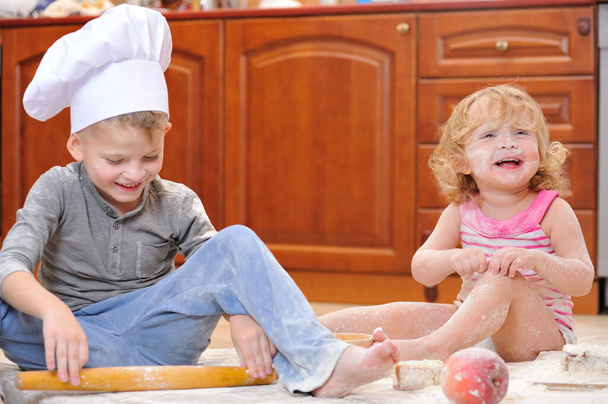 Két testvér - fiú és lány - séf sapkában a kandalló mellett ül a konyhapadlón liszttel beszennyezve, ételekkel játszva, rendetlenséget csinálva és szórakozva. - Fotó, kép
