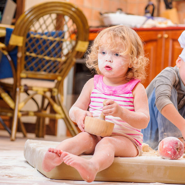 Δύο αδέλφια - αγόρι και κορίτσι - με καπέλα σεφ κοντά στο τζάκι κάθονται στο πάτωμα της κουζίνας λερωμένα με αλεύρι, παίζοντας με το φαγητό, κάνοντας χάος και διασκεδάζοντας - Φωτογραφία, εικόνα