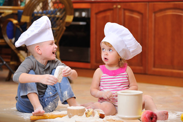 Dva sourozenci - chlapec a dívka - v kuchařských čepicích u krbu, sedící na podlaze v kuchyni poskvrněné moukou, hrající si s jídlem, dělají nepořádek a baví se - Fotografie, Obrázek