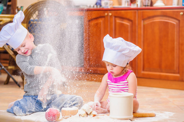 Two siblings - boy and girl - in chef's hats near the fireplace sitting on the kitchen floor soiled with flour, playing with food, making mess and having fun - Photo, Image