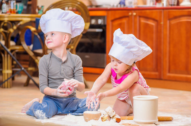 Dos hermanos - niño y niña - en sombreros de chef cerca de la chimenea sentados en el suelo de la cocina manchados de harina, jugando con la comida, haciendo lío y divirtiéndose - Foto, Imagen