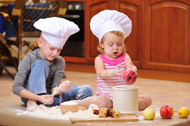 Dos hermanos - niño y niña - en sombreros de chef cerca de la chimenea sentados en el suelo de la cocina manchados de harina, jugando con la comida, haciendo lío y divirtiéndose - Foto, imagen