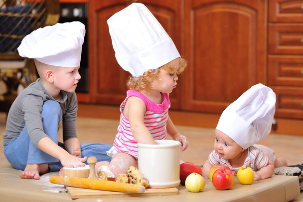 兄弟-男の子と女の子-シェフの帽子の中で暖炉の近くに小麦粉で汚れたキッチンフロアに座って、食べ物で遊んで、混乱を作り、楽しんで - 写真・画像