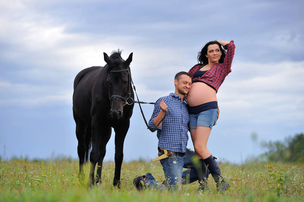 Joyeux couple - elle est belle brune aux cheveux longs, enceinte ; il est à genoux, embrassant son ventre et tenant les rênes du cheval noir. prairie, ciel merveilleux derrière - Photo, image