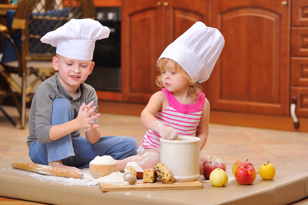 Deux frères et sœurs - garçon et fille - dans les chapeaux du chef près de la cheminée assis sur le sol de la cuisine souillé avec de la farine, jouer avec la nourriture, faire du désordre et s'amuser - Photo, image